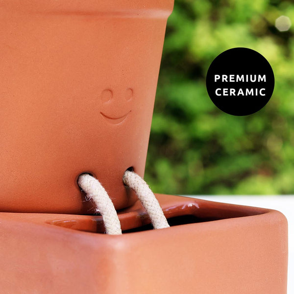 Terracotta pot with a smiley face design and rope legs, labelled 'Premium Ceramic' against a green garden background.