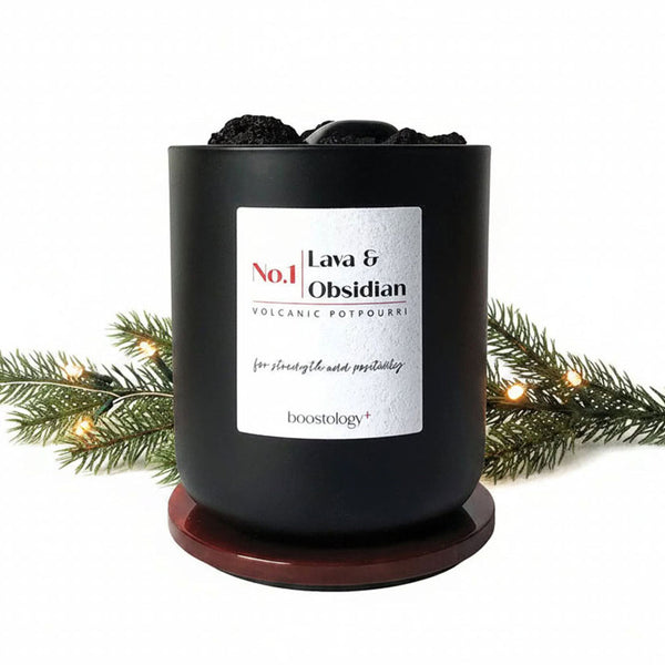 A matte black volcanic potpourri jar with a white label reading 'No.1 Lava & Obsidian' and the Boostology brand name, styled with a green Christmas tree branch and warm fairy lights on a white background.