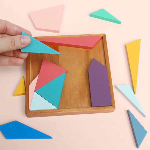 Colourful geometric shapes in a wooden tray with a hand arranging the pieces, on a light pink background.