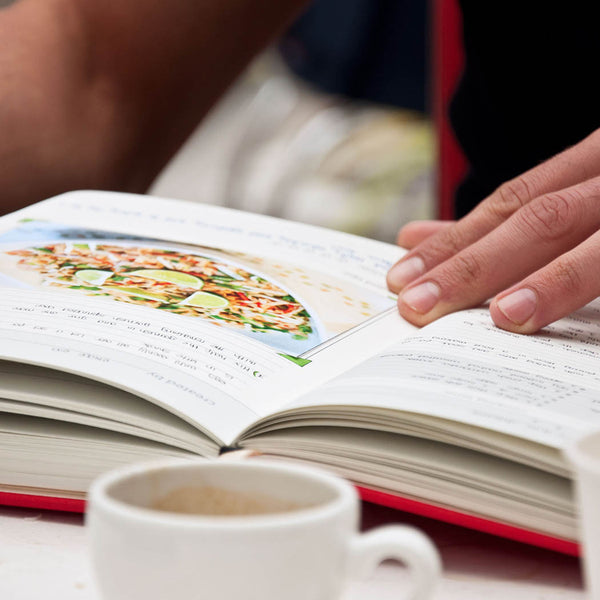 Open cookbook with a colorful image of a dish, a hand holding the page, and a cup of coffee on a table.