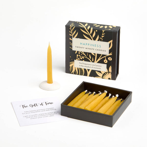 An opened box of yellow beeswax candles displayed on a white background. The lid reads 'HAPPINESS TWENTY MINUTE CANDLES' The photo also shows a single candle in a small white holder, and a printed card titled 'The Gift of Time'.