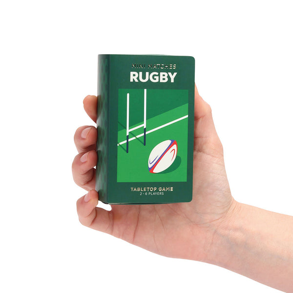 A hand holding a green rugby tabletop game box against a plain white background. The box has white text reading 'MINI MATCHES RUGBY' and 'TABLETOP GAME 2-6 PLAYERS' with a simple rugby ball and field graphic on the front.