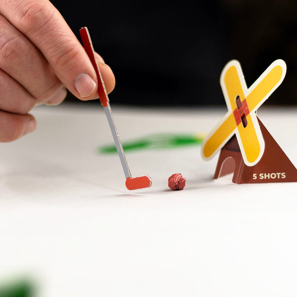 Hand holding a small golf club near a miniature windmill on a white surface.