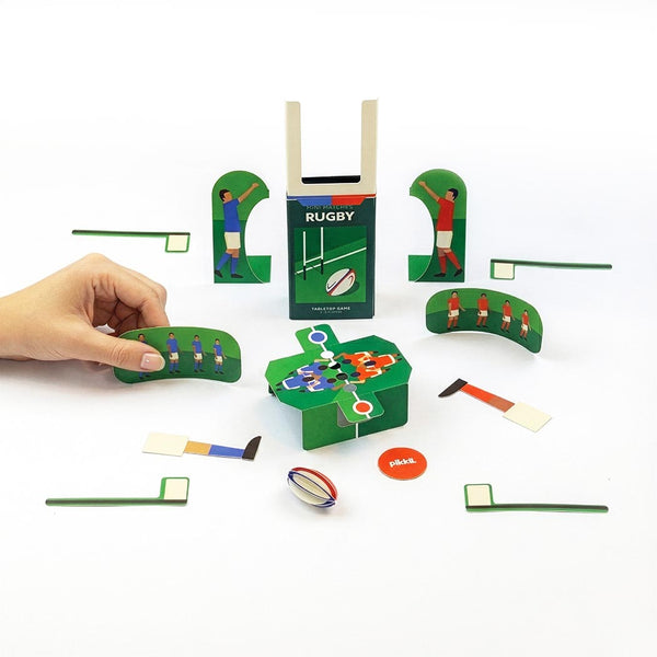 A tabletop rugby game set displayed on a white background, including goal post, players, pitch markings and a hand holding one of the curved playing pieces. 