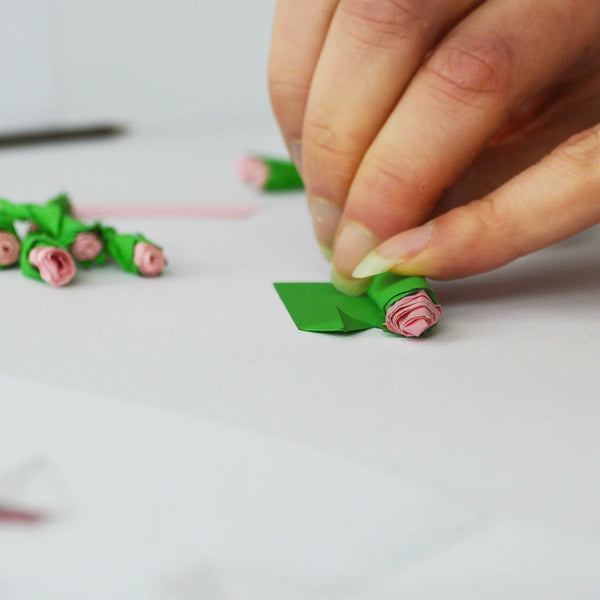 Hand making paper flowers with green stems and pink petals on a white surface.
