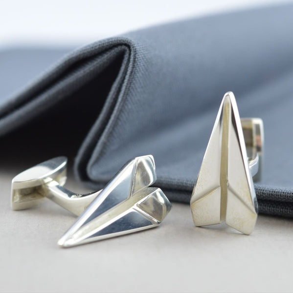 Two Recycled Silver paper plane cufflinks lying next to a grey shirt